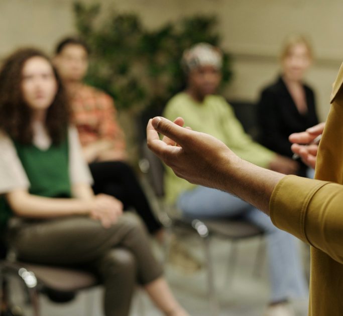 Person spricht vor einer Gruppe von fünf Menschen in einem Sitzungsaal.