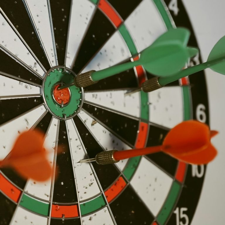 Dartboard mit drei Pfeilen, zwei in Rot und einer in Grün, die im Ziel stecken.