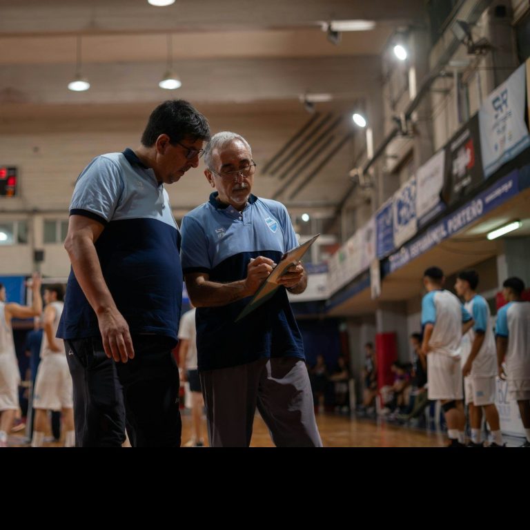 Zwei Trainer besprechen Taktiken am Basketballspielfeld. Spieler im Hintergrund aktiv.