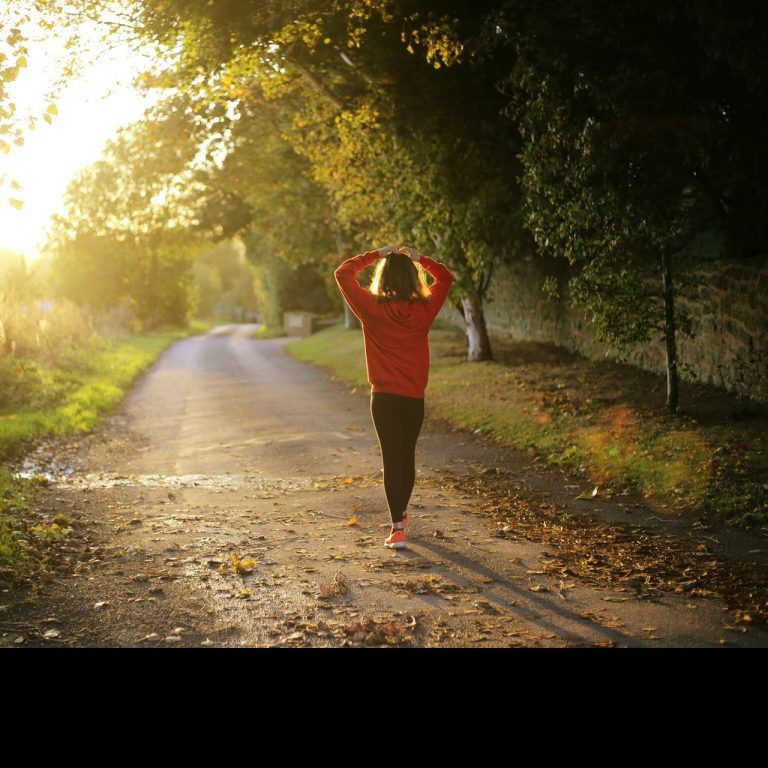 Eine joggende Person in roter Jacke auf einem Sonnenlicht durchfluteten Weg.