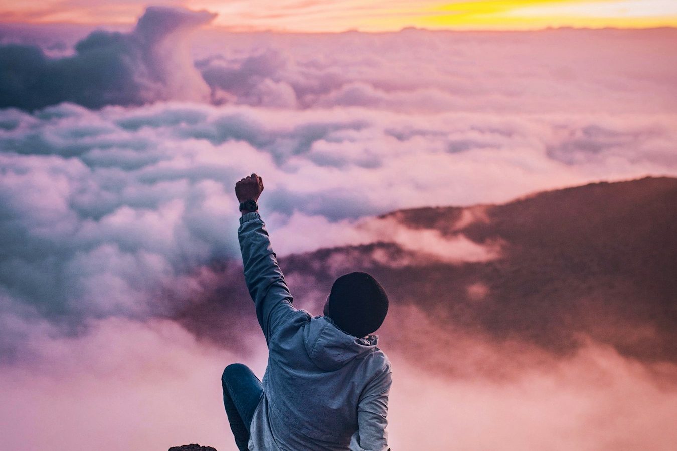 Mann sitzt auf einem Berg und hebt die Faust triumphierend gegen einen bunten Sonnenuntergang.